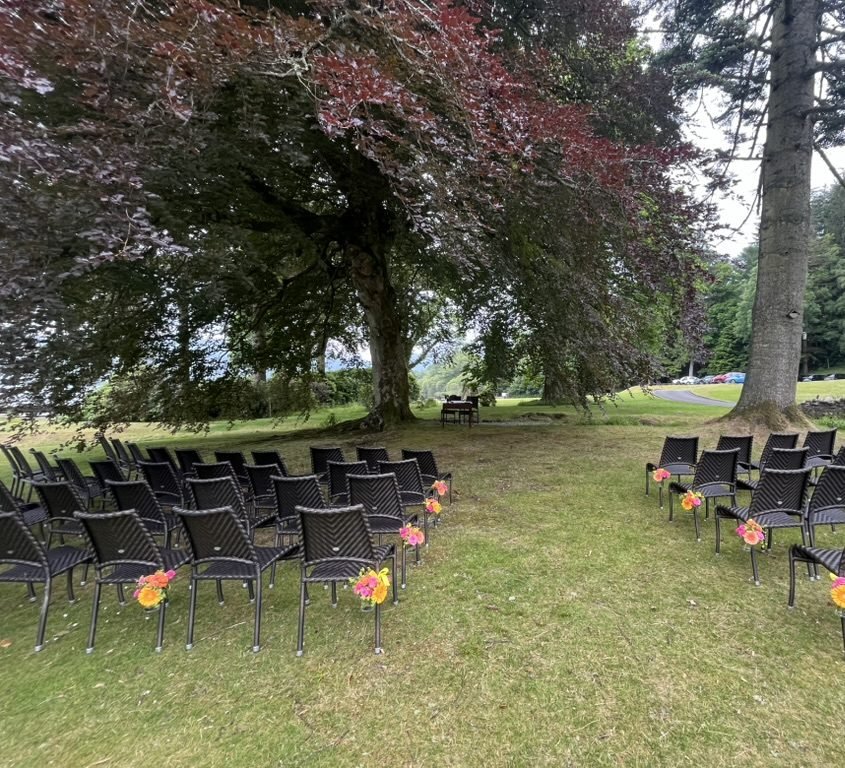 Rows of dark wicker chairs on the lawn at Altskeith Country House, bright orange and pink flowers tied to the aisle ends, set out under a huge copper beech with a small signing table beneath it