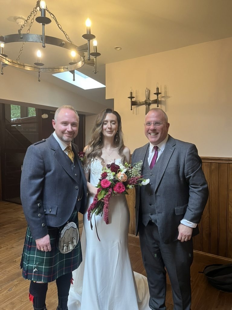 Andrew in a kilt and Alicia in a fitted white wedding gown with her bouquet, standing with celebrant John Paul Fitzpatrick in a wood-panelled room at Crossbasket Castle