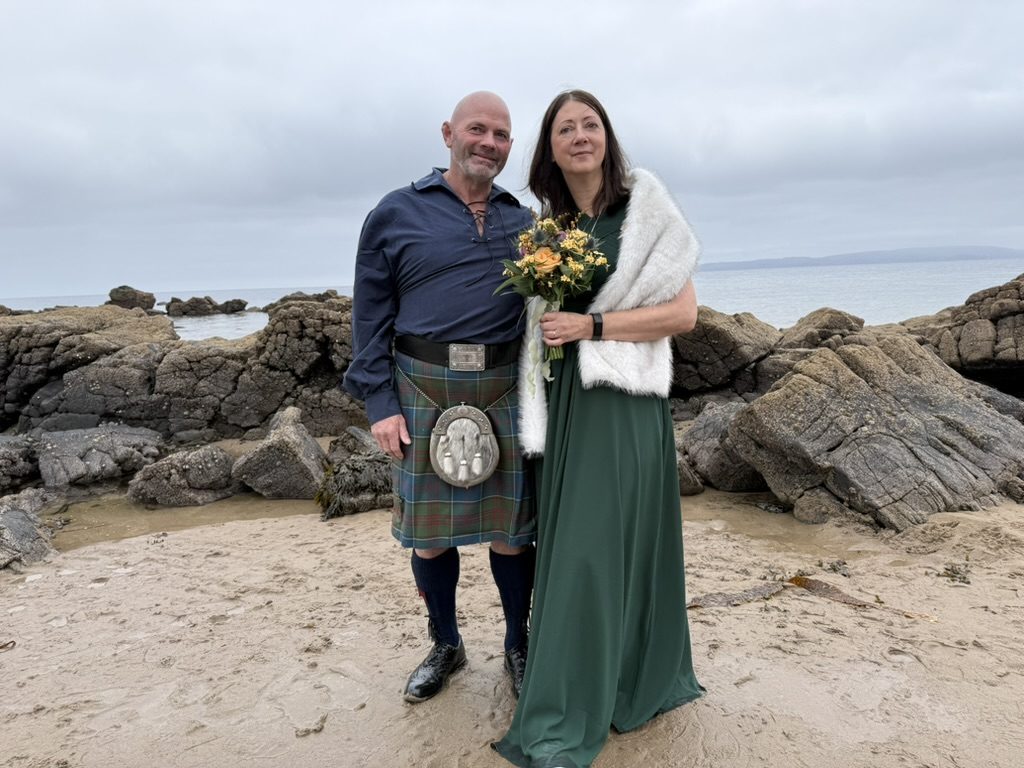 A couple celebrating their humanist wedding on an Arran beach, sea and rocks behind