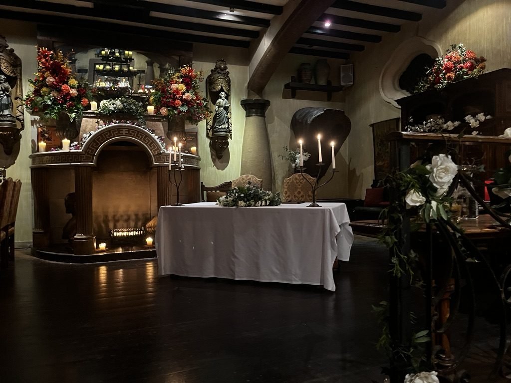 The fireplace at Artà set for a humanist wedding: a signing table in white linen with a garland of greenery and two lit iron candelabras, in front of a carved fireplace crowned with red and orange flowers, under dark timber beams