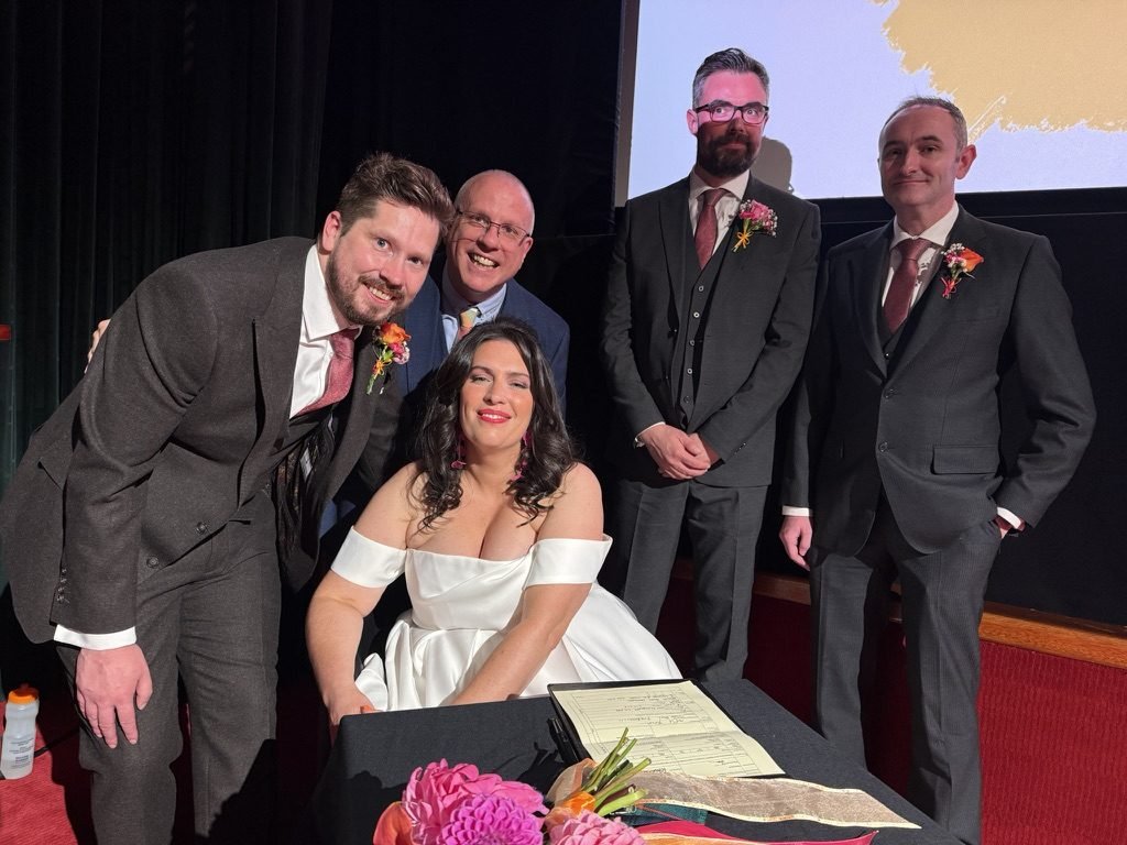 Becky and Tom signing the marriage schedule at a table in front of the cinema screen, their witnesses standing behind them