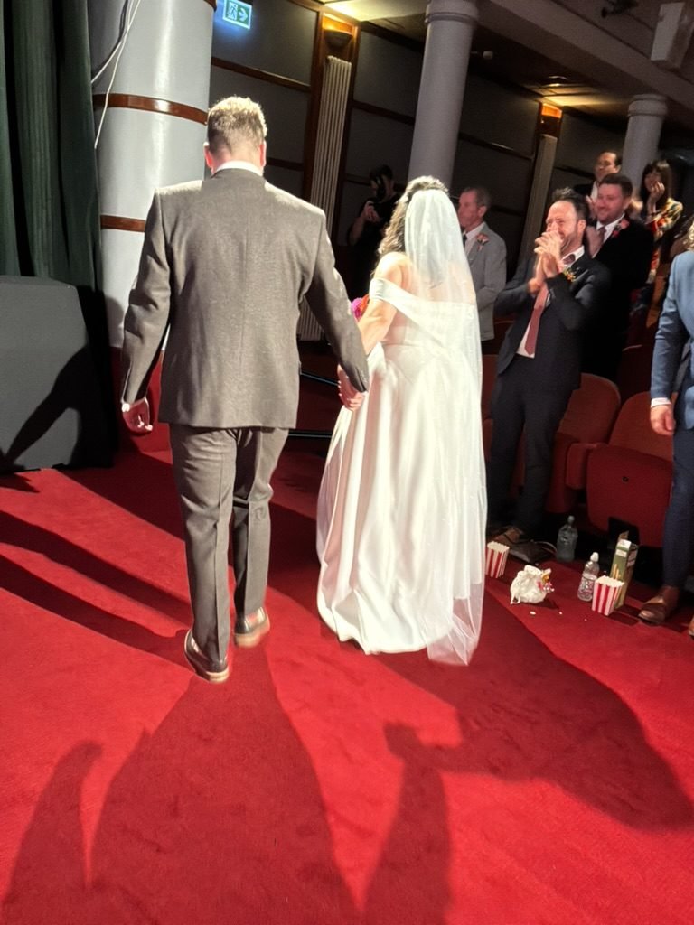 Becky and Tom walking hand in hand back up the red carpet aisle of the cinema after their ceremony, guests applauding from the seats