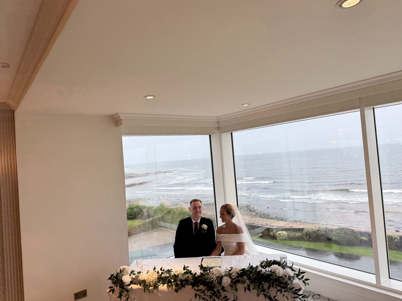 Ben and Jessica sitting at the signing table in the corner window of the Firth Pavilion at Seamill Hydro, white flowers along the table and the Firth of Clyde and the shoreline behind them