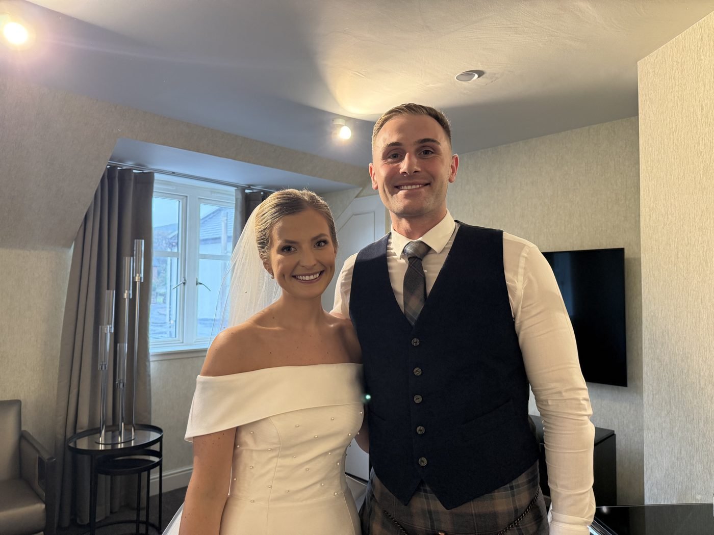 Jessica in an off-the-shoulder gown and veil and Ben in a navy waistcoat and kilt smiling together in a room at Seamill Hydro