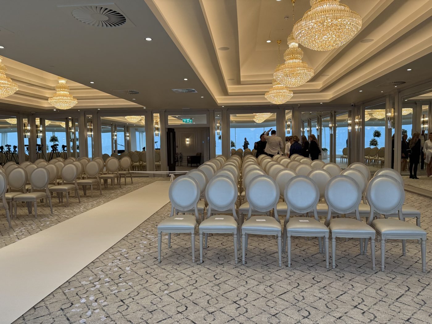 The Firth Pavilion at Seamill Hydro set for the ceremony, rows of white upholstered chairs either side of a white aisle beneath crystal chandeliers, with a wall of windows onto the sea