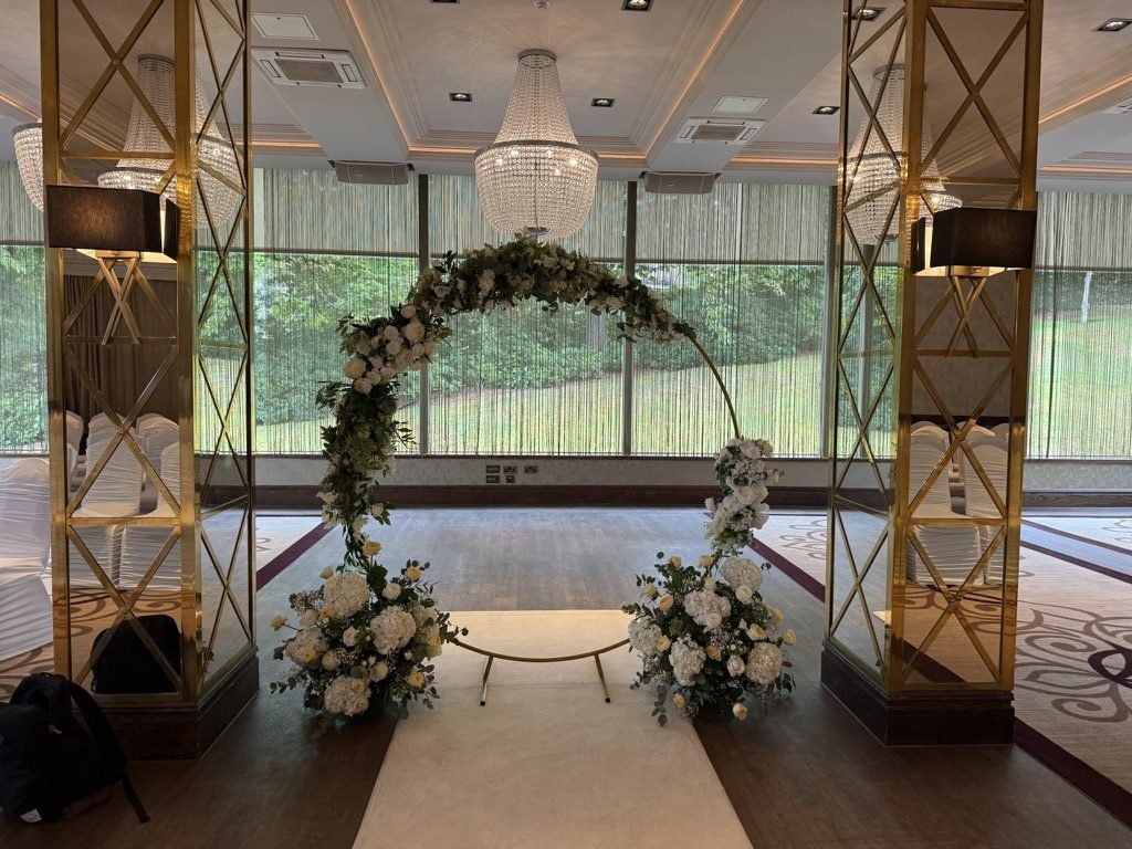 The Grand Ballroom at Boclair House set for a humanist ceremony in July 2025: a round flower arch on a cream aisle runner, framed by mirrored gold pillars and crystal chandeliers, with tall windows onto the lawn and trees behind