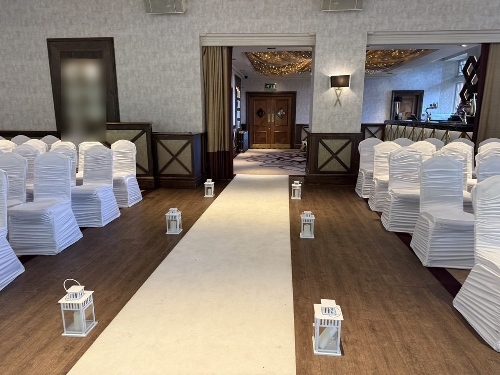 Looking back down the aisle of the Boclair House ballroom as it was in July 2025: white lanterns along a cream runner, rows of white-covered chairs on a dark wooden floor, and wooden double doors at the far end