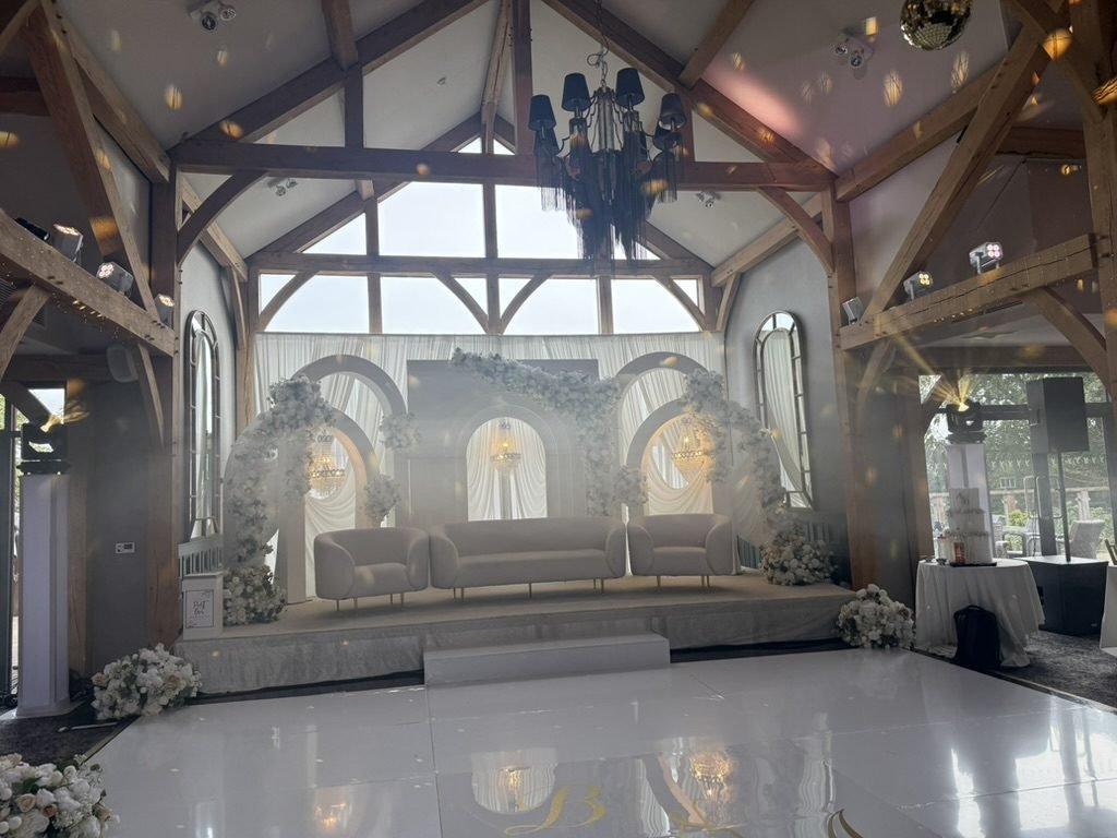 The function room at Enterkine House dressed for Brycen and Ammara's wedding celebration, oak roof beams and a tall glazed gable above a white floral stage with cream sofas, chandeliers and a gloss white dance floor
