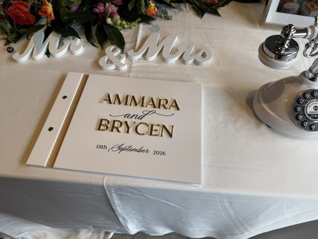 A white guest book with gold lettering reading Ammara and Brycen, 13th September 2026, on a white-clothed table at Enterkine House beside wooden Mr and Mrs letters, bright flowers and a vintage white telephone