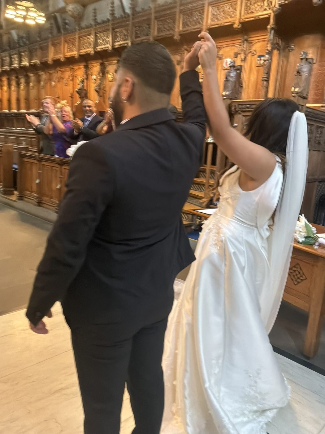 Burak and Duygu walking back down the aisle of the University of Glasgow Memorial Chapel with their hands raised together, guests applauding beside the carved wooden stalls