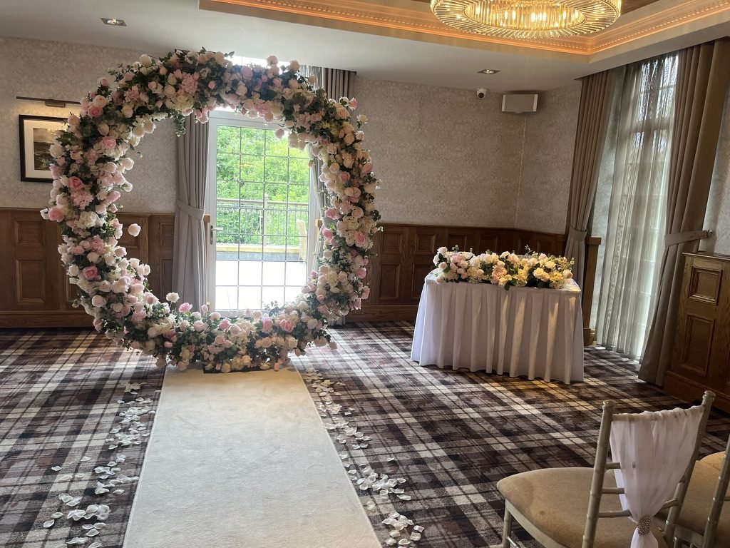 The Busby Hotel ceremony room set for a humanist wedding in June 2024: a round arch of pink and white flowers on a cream aisle runner, a signing table beside a glazed door to the balcony, tartan carpet and wood panelling