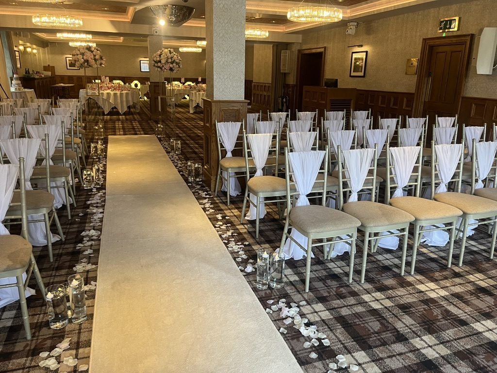 Looking down the cream aisle runner at The Busby Hotel: gold chairs with white sashes either side, candles and petals along the aisle, and the dining tables set at the far end of the room