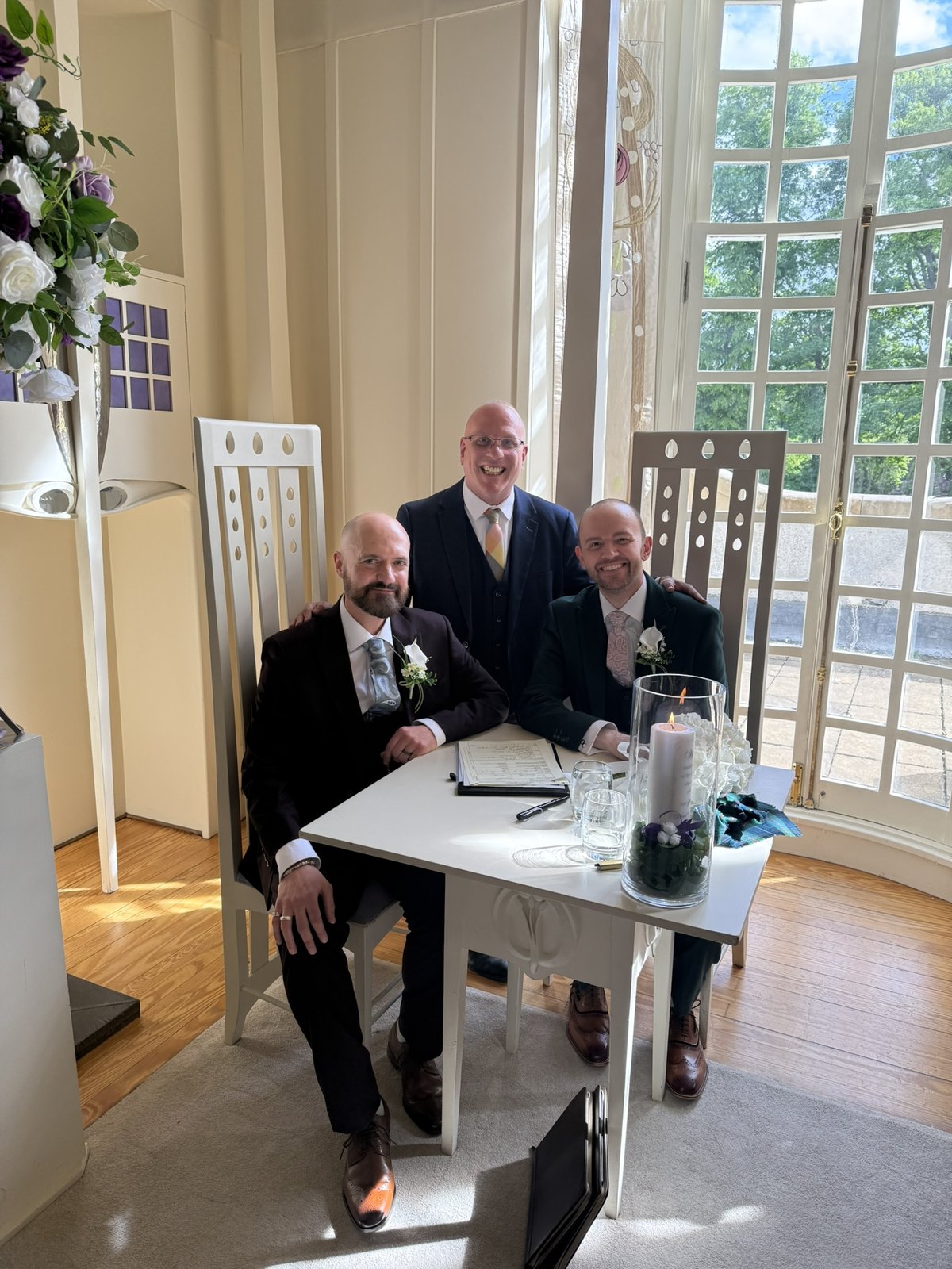 Chris and David with celebrant John Paul at the signing table at House for an Art Lover, Glasgow — two grooms and their Marriage Schedule