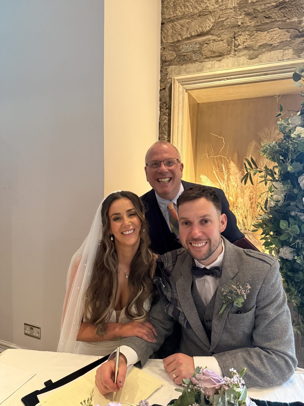 Claire and Russell smiling at the signing table at Citation with celebrant John Paul leaning in behind them, Russell holding the pen over the marriage schedule, lilac roses on the table and a sandstone wall behind