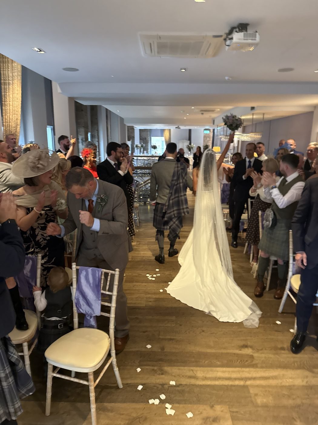Claire punching her bouquet into the air as she and Russell, in his kilt and grey tweed jacket, head back up the aisle at Citation after the ceremony, guests on their feet clapping and petals scattered on the wooden floor