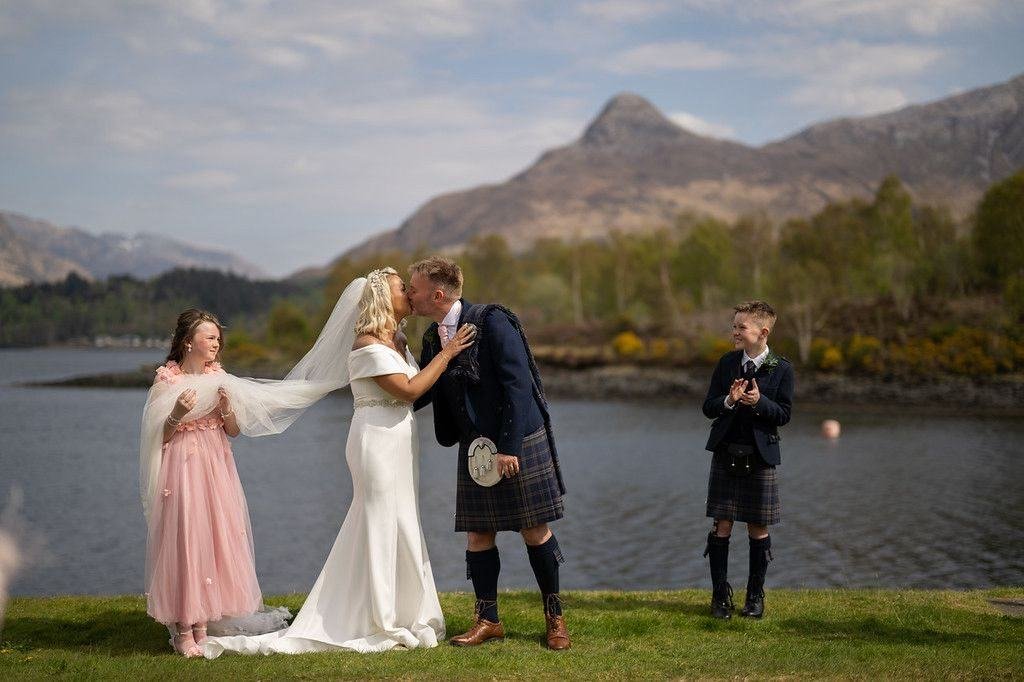 Colin and Lisa Marie share their first kiss as a married couple on the shore of Loch Leven at the Isles of Glencoe, their two children beside them and the Pap of Glencoe behind