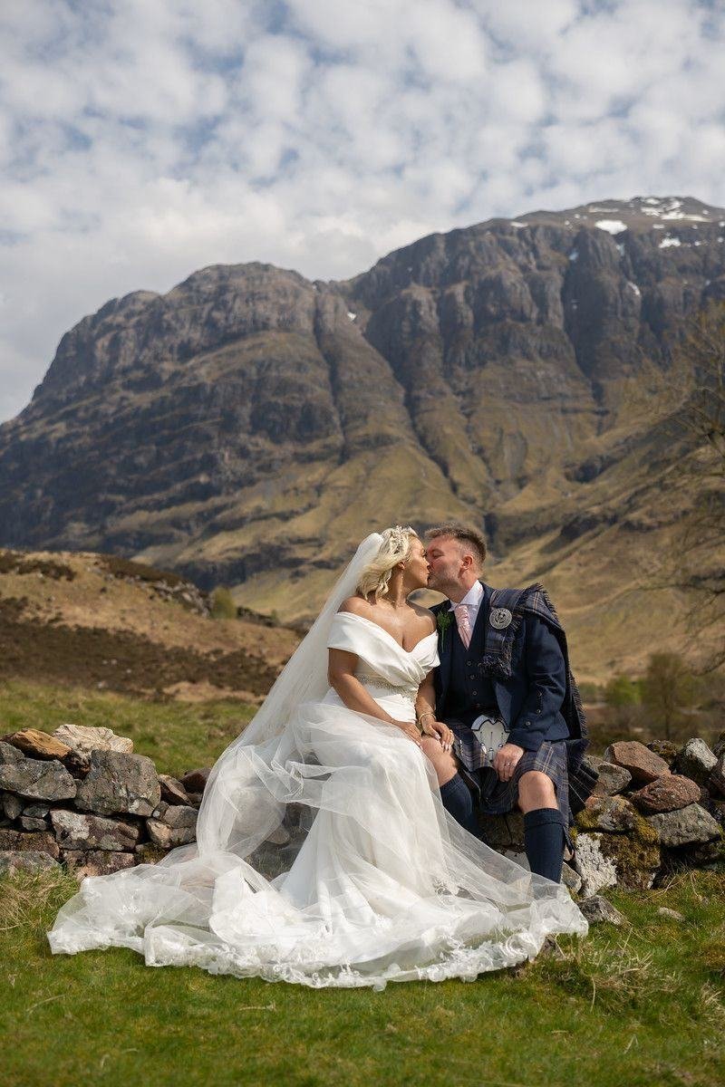 Colin and Lisa Marie kiss on a dry-stone wall in Glencoe, the mountains rising behind them