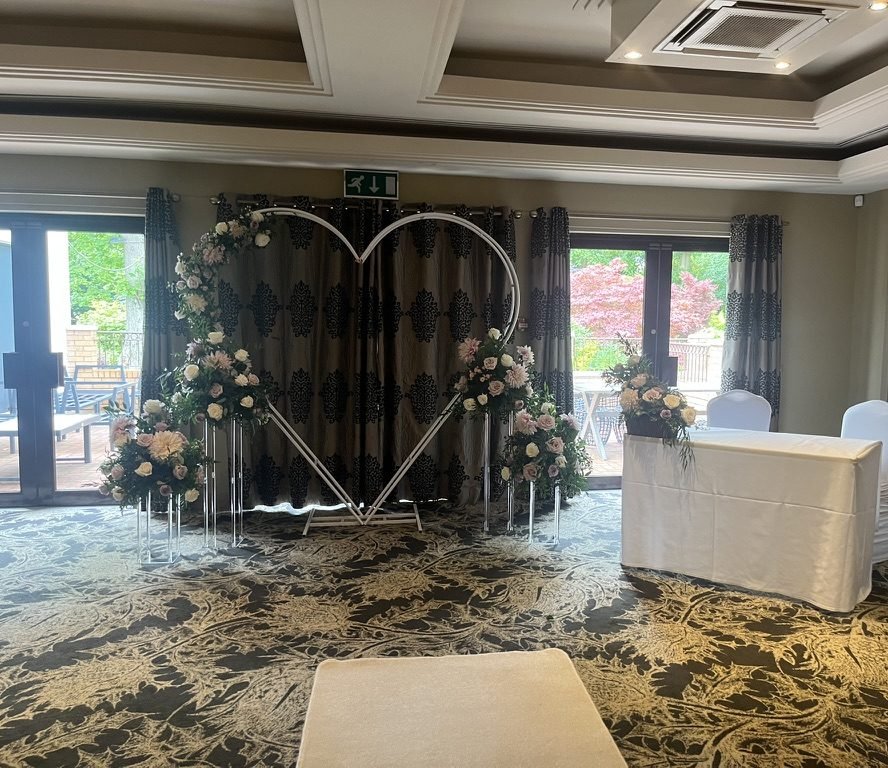 A function suite at Dalziel Park Hotel set for a humanist wedding in July 2024: a white heart-shaped arch dressed with roses in front of patterned curtains, a signing table to one side, and French doors onto a patio and a red-leaved tree