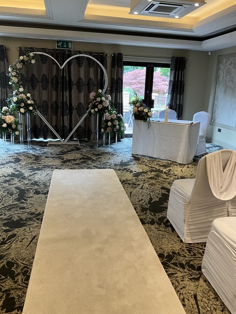 The cream aisle at Dalziel Park Hotel leading to a white heart-shaped arch and the signing table, white-covered chairs to one side and a red-leaved tree through the French doors behind