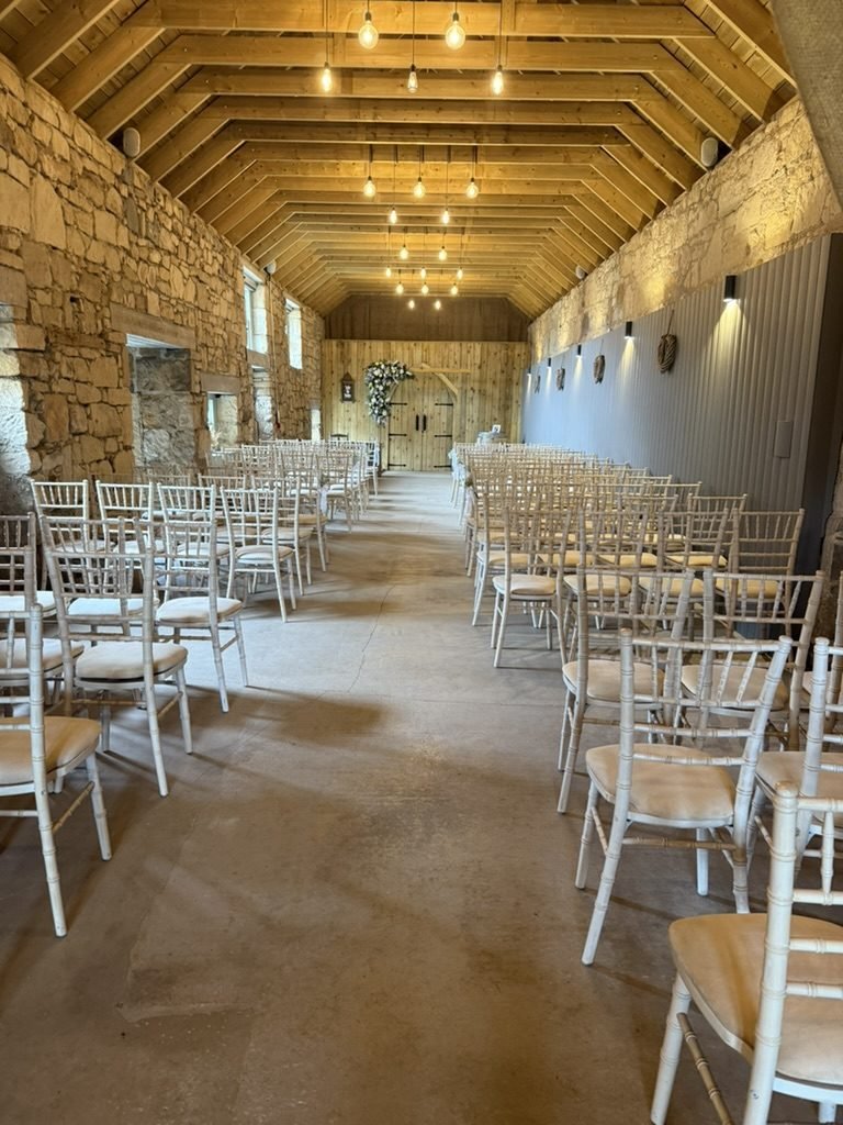 The Byre at The Den at Culross set for a humanist wedding, rows of white chairs down a long room between a sandstone wall and grey panelling, bulbs hanging from exposed timber trusses, and arched timber doors with a spray of white flowers at the far end