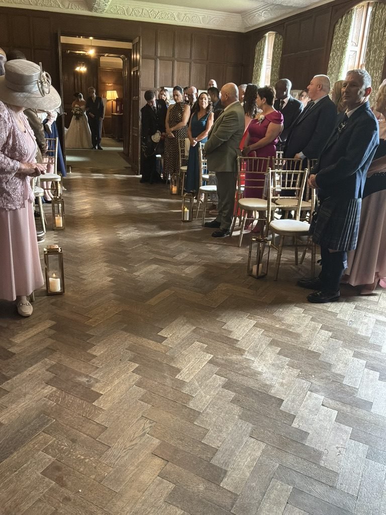 Egle waiting at the doorway of the panelled Ceremony Room at Rowallan Castle, lanterns and candles lining the aisle, guests standing on both sides