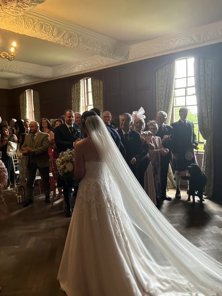 Egle in her dress with a long veil in the panelled Ceremony Room at Rowallan Castle, guests applauding beneath the ornate plasterwork ceiling, Cookie the dog watching from the side
