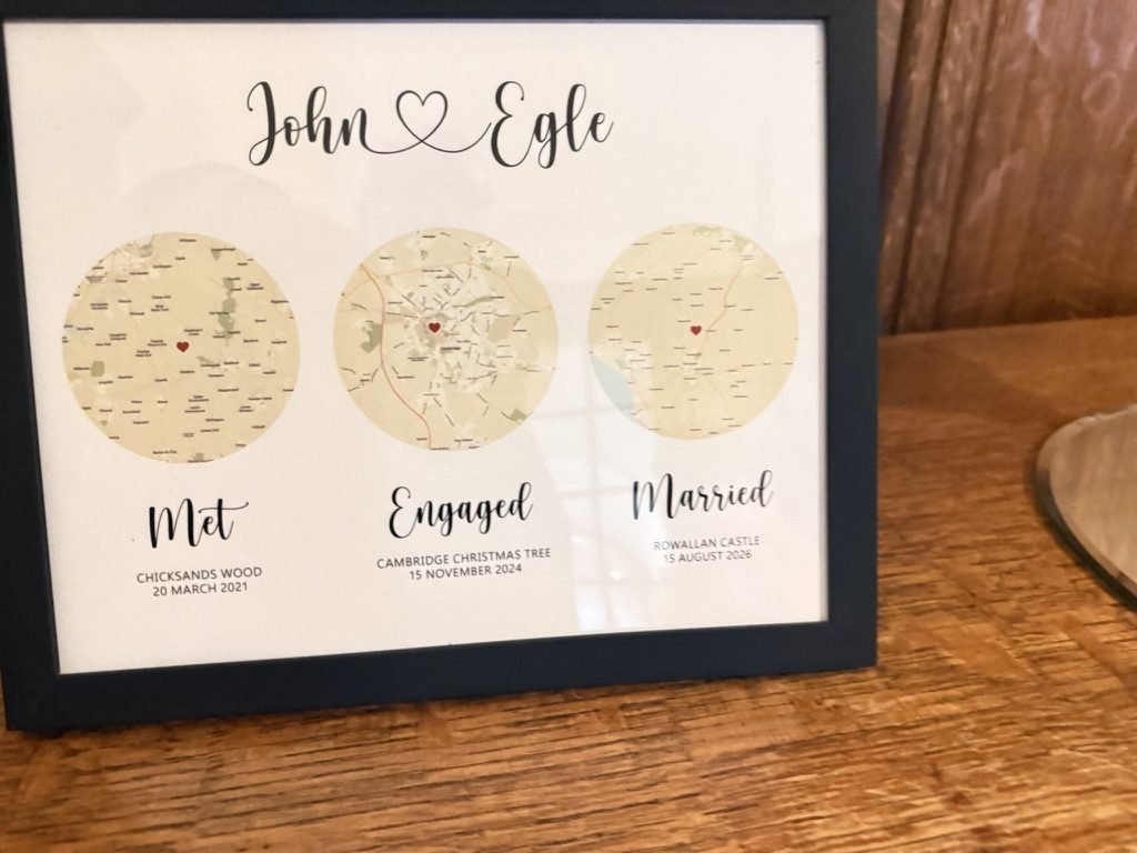 A framed map print displayed at the wedding marking where Egle and John met, got engaged and married, resting on an oak table