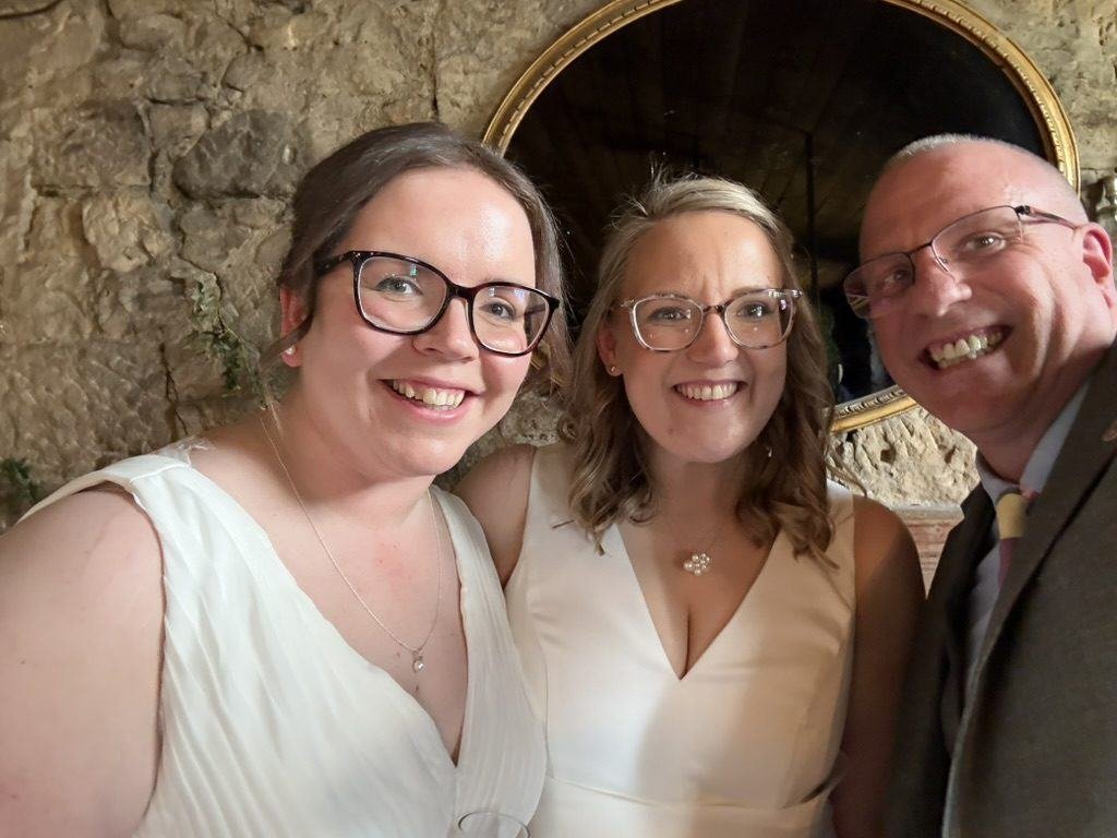 Emily and Gill with celebrant John Paul in front of the stone wall and mirror at The Bothy