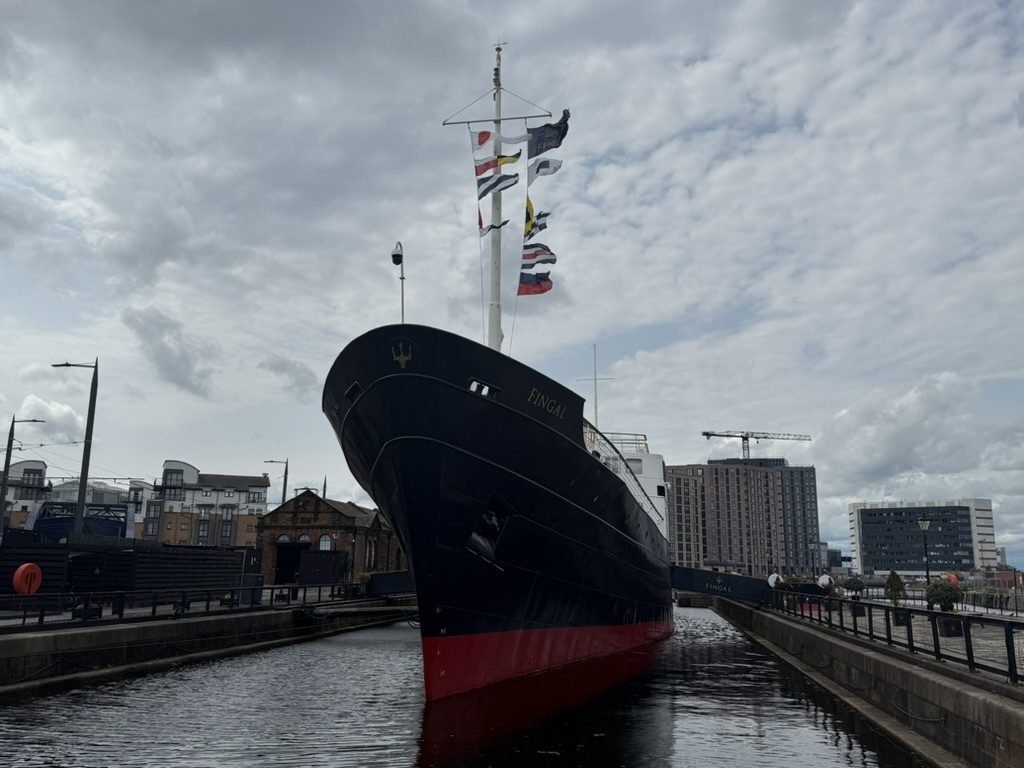 The bow of Fingal at her berth in Leith, black hull and red waterline reflected in the dock, a string of signal flags running up the mast under a grey sky