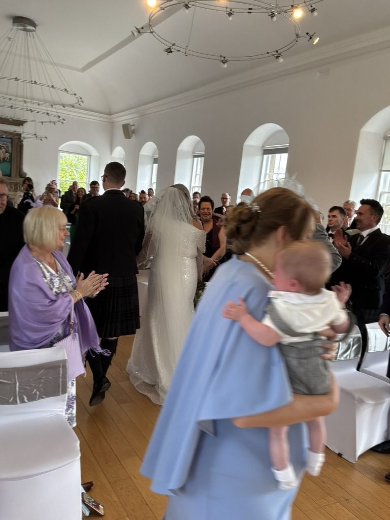 Frances and Kevin walking back down the aisle at West Lothian Burgh Halls after their wedding ceremony, guests applauding on both sides
