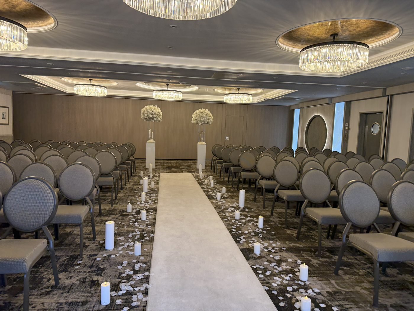 The Lagavulin Suite at the Gailes Hotel set for a humanist wedding, rows of grey chairs either side of a long white aisle lined with candles and petals, a white flower arch in front of the windows and chandeliers overhead