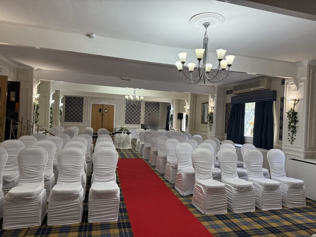 The ceremony room at the Glazert Country House Hotel set for a humanist wedding in July 2025: a red carpet aisle between rows of white-covered chairs, tartan carpet, chandeliers overhead and a small signing table at the far end