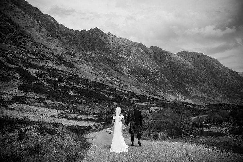 Colin and Lisa Marie walk hand in hand down the road through Glen Coe after their wedding, the Aonach Eagach ridge towering above them, in black and white