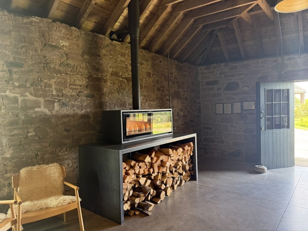 Inside the old steading at Guardswell Farm: rough stone walls under a pitched timber roof, a wood stove set above a long stack of split logs, and a green stable door open to the daylight