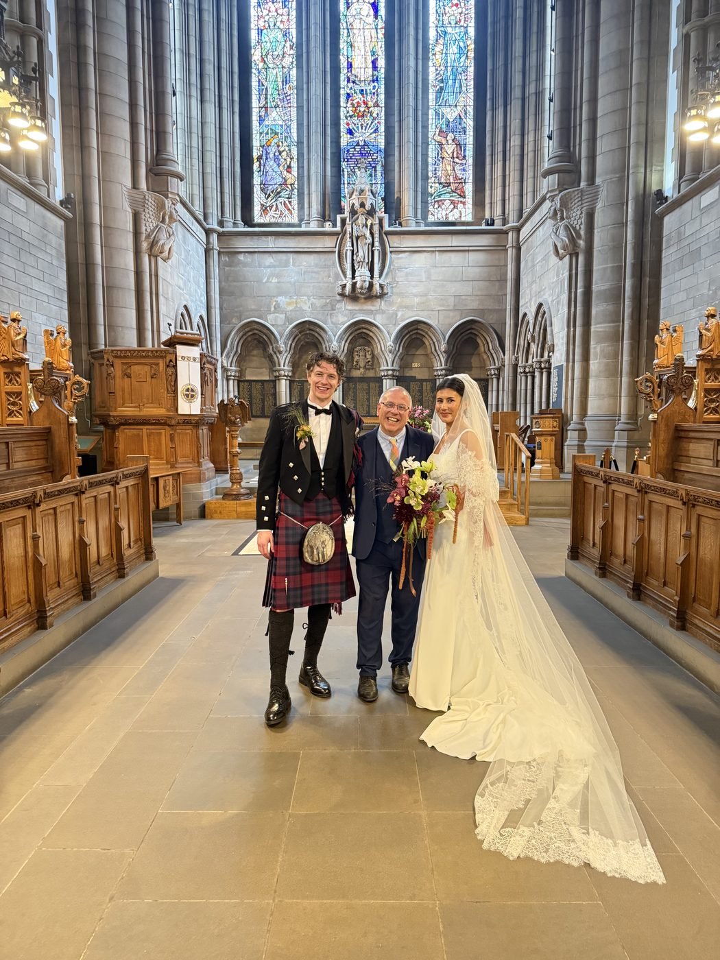 Hannah in a long lace-edged veil and Euan in a kilt and black jacket standing with celebrant John Paul in the aisle of Glasgow University Chapel, carved wooden stalls on either side and tall stained glass windows behind