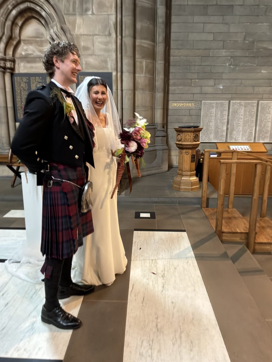 Euan in his kilt and Hannah in her veil laughing together at the front of Glasgow University Chapel, war memorial panels on the stone wall behind them