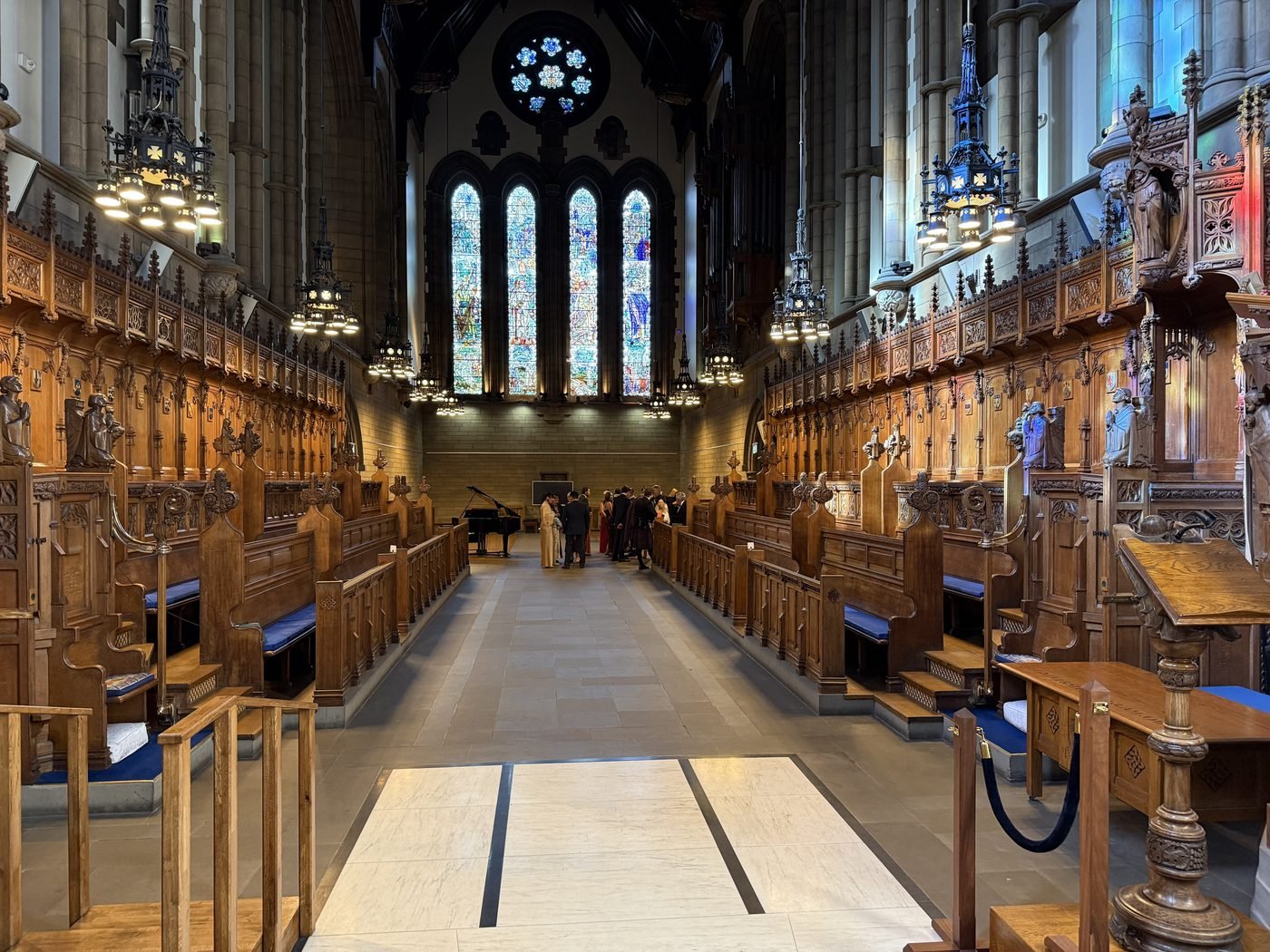 The long central aisle of Glasgow University Chapel, lined with carved oak stalls and blue cushions, running towards Douglas Strachan's stained glass windows and rose window