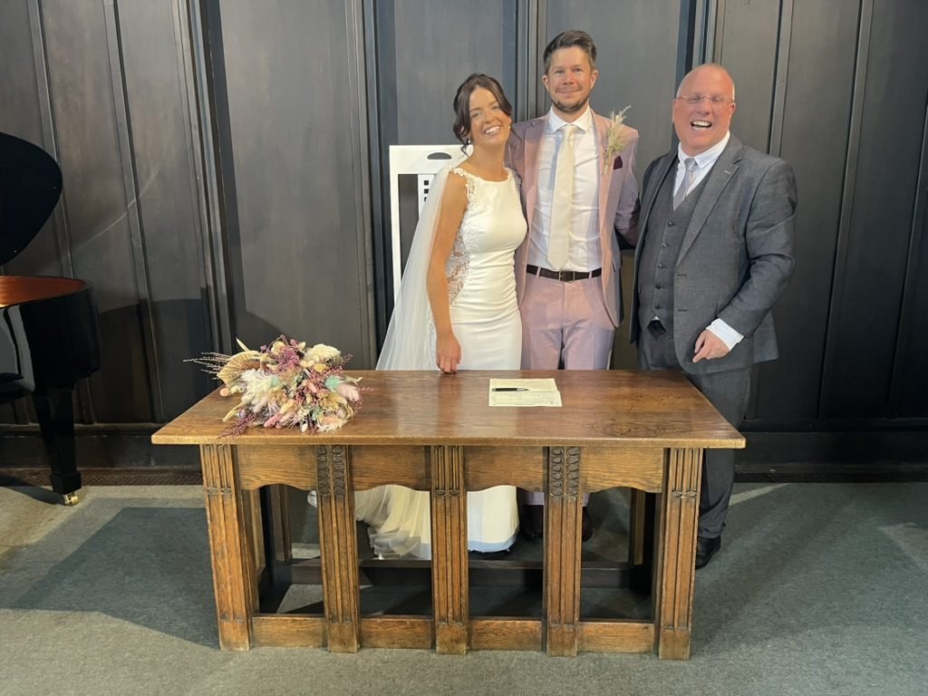 Hazel and Matt standing behind the carved oak signing table in the Mackintosh Church with celebrant John Paul beside them, dark timber panelling behind, a grand piano to one side and a dried-flower bouquet on the table