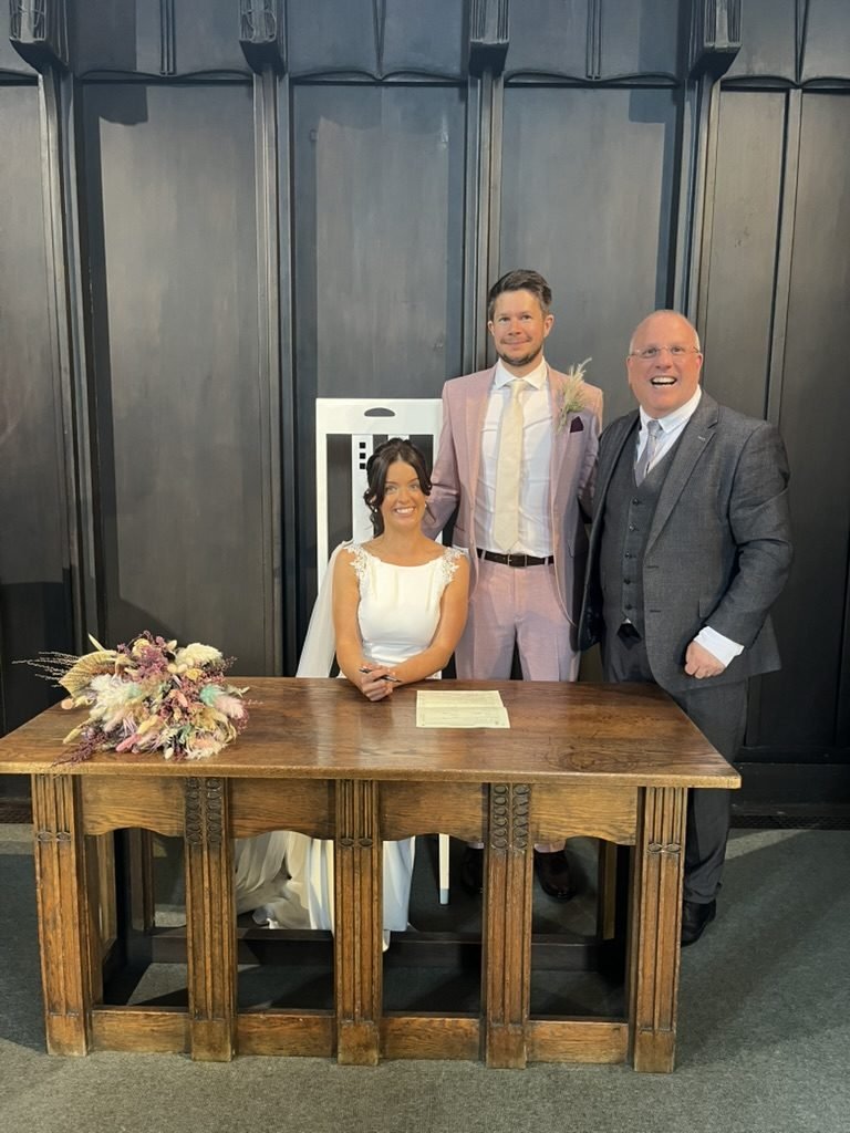 Hazel seated at the carved oak signing table in the Mackintosh Church, pen in hand, with Matt standing behind her in a pink suit and celebrant John Paul beside them, a Mackintosh high-backed chair behind