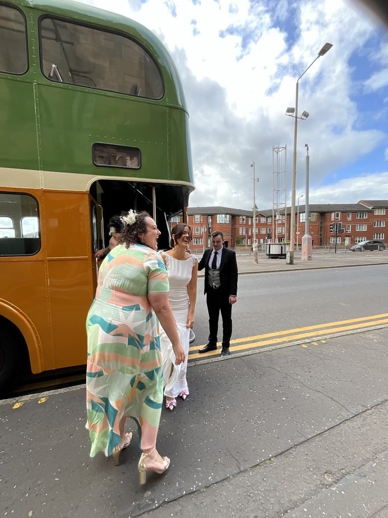 Hazel in her wedding dress laughing with a friend beside a vintage green and cream Glasgow double-decker bus, red sandstone tenements and a blue sky behind