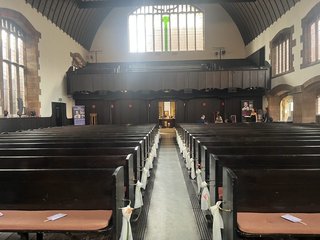 The inside of the Mackintosh Church at Queen's Cross dressed for Hazel and Matt's wedding, dark timber pews with white ribbons and pink calla lilies along the aisle, tall leaded windows and the timber gallery at the far end