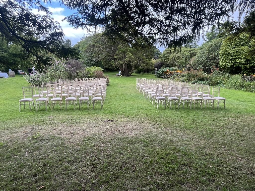 Rows of pale wooden chairs set out in two blocks on the lawn of the Plaisance, the walled garden at Kelburn, under the spreading branches of old conifers, with flowering shrubs and borders beyond