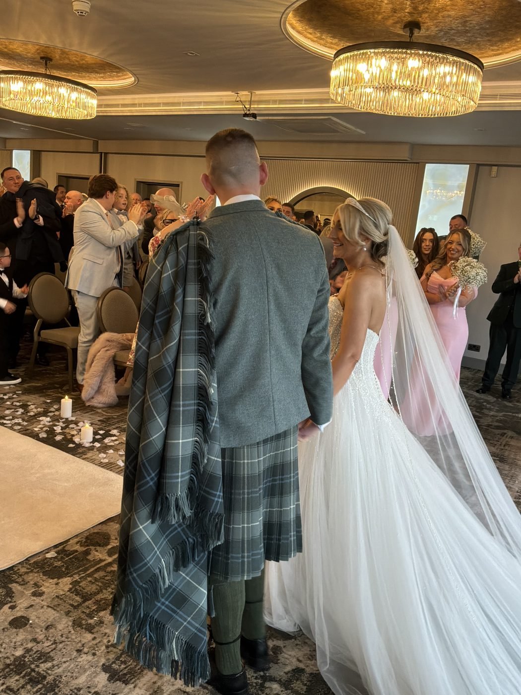 Michael in a grey tweed jacket and kilt with his tartan plaid over his shoulder, beside Kelly in a strapless gown and long veil, as guests stand and applaud under the chandeliers of the Lagavulin Suite at the Gailes Hotel