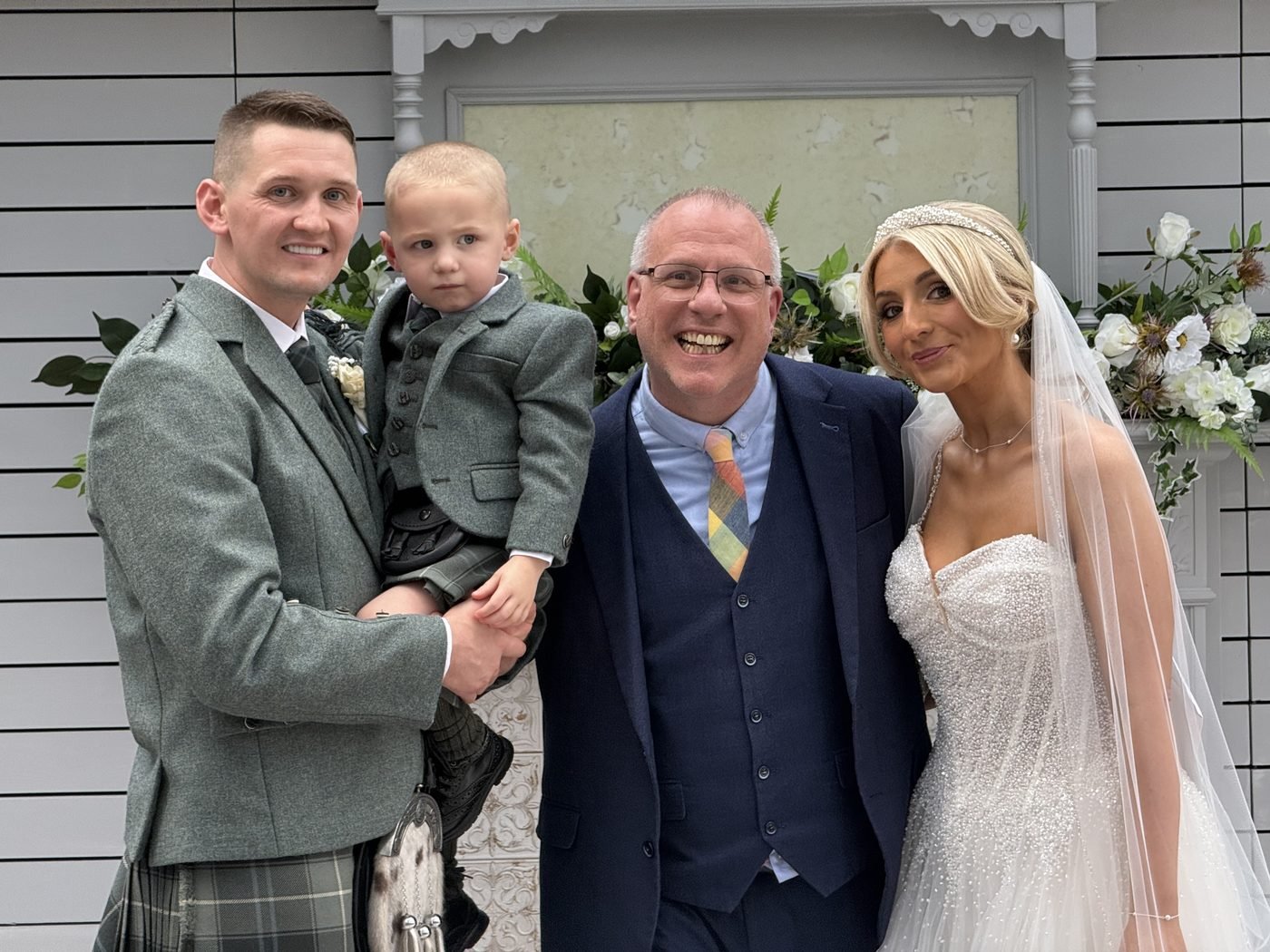 Michael holding their young son in a matching grey jacket, celebrant John Paul in a navy suit, and Kelly in her veil, all smiling in the Rooftop Conservatory at the Gailes Hotel