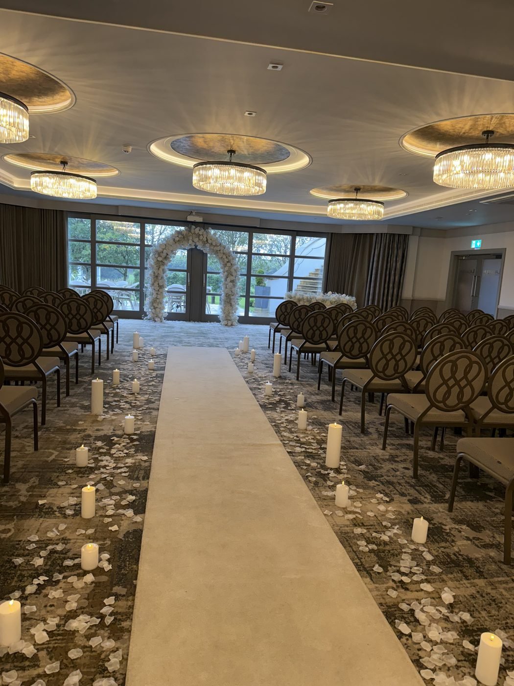 The Lagavulin Suite at the Gailes Hotel set for the ceremony, a long white aisle lined with candles and scattered petals between rows of grey chairs, leading to a white flower arch in front of tall windows, with chandeliers overhead