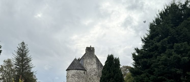 The grey stone tower of Kilmartin Castle, with its round turret and conical slate roof, rising between tall conifers under a heavy Argyll sky