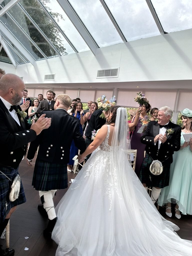 Kyle and Kirsty walking hand in hand back down the aisle after their ceremony at Sherbrooke Castle Hotel, guests applauding on both sides beneath the glass roof