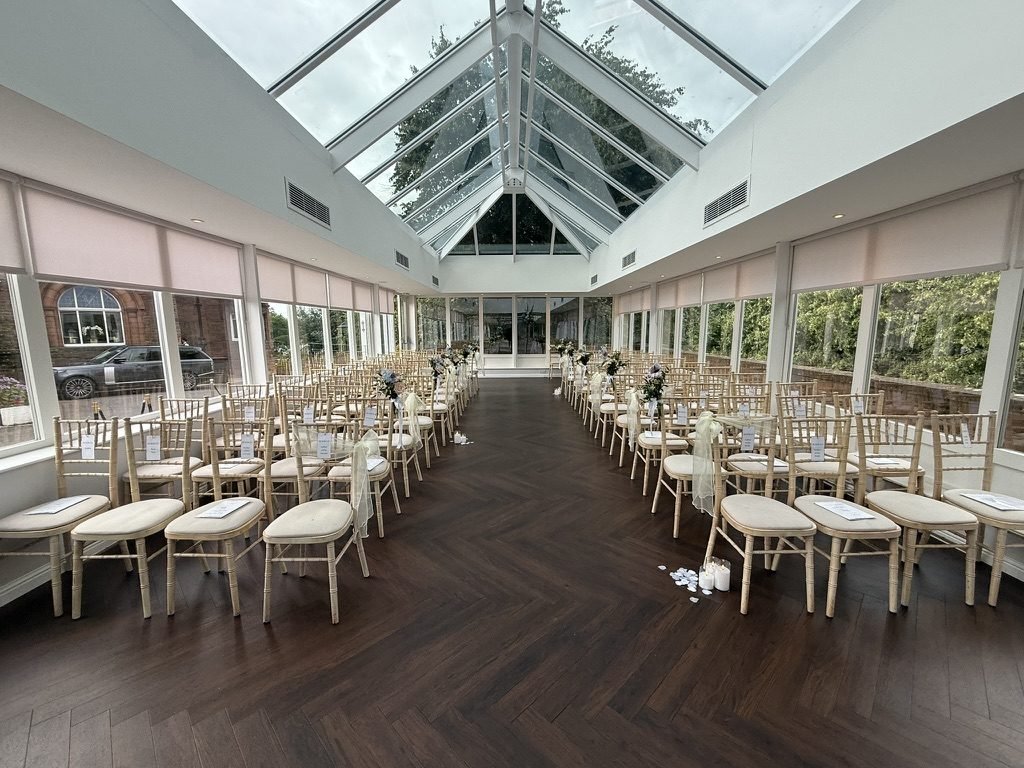 The Orangery at Sherbrooke Castle Hotel set for a wedding ceremony, chiavari chairs either side of a herringbone floor aisle beneath a pitched glass roof