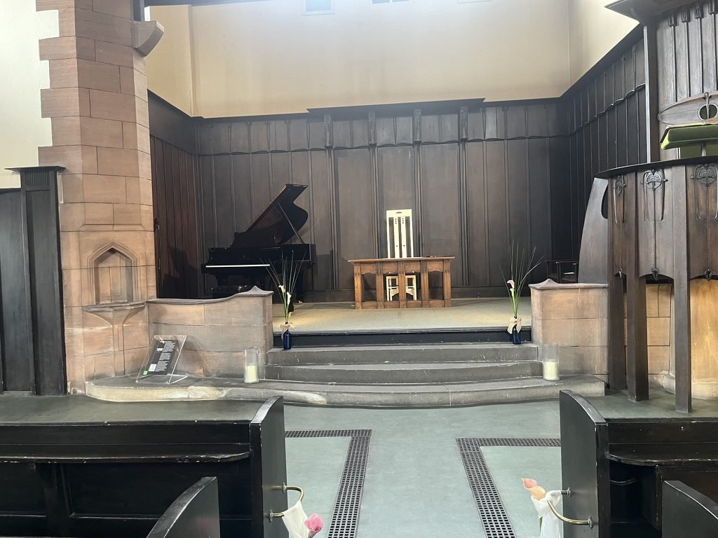 The chancel of the Mackintosh Church at Queen's Cross ready for a wedding: a carved oak table and a high-backed Mackintosh chair in front of dark timber panelling, a grand piano to one side and calla lilies at the foot of the sandstone steps