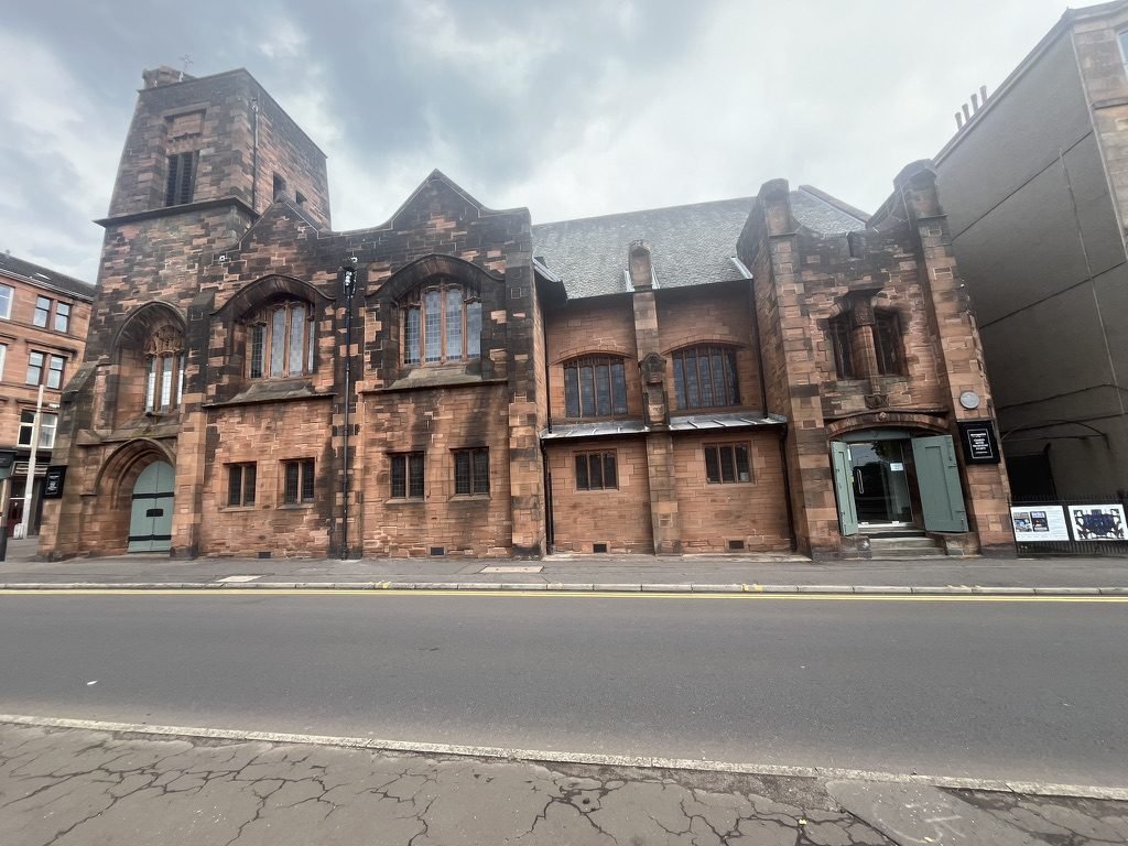 The red sandstone front of Mackintosh Queen's Cross on Garscube Road in Maryhill, a square tower at one end and an open door under a carved arch at the other, beneath a grey Glasgow sky
